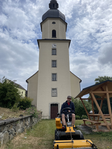 Mann auf Rasentraktor mäht Gras vor einer KIrche - Kustus, Küster, Kircher, Mesner und Co.