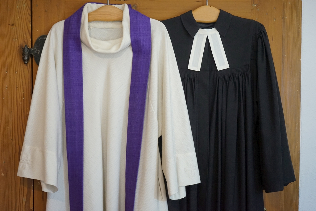 Kirchengewänder: weiße Alba mit lila Stole neben schwarzen Talar mit weißen Beffchen